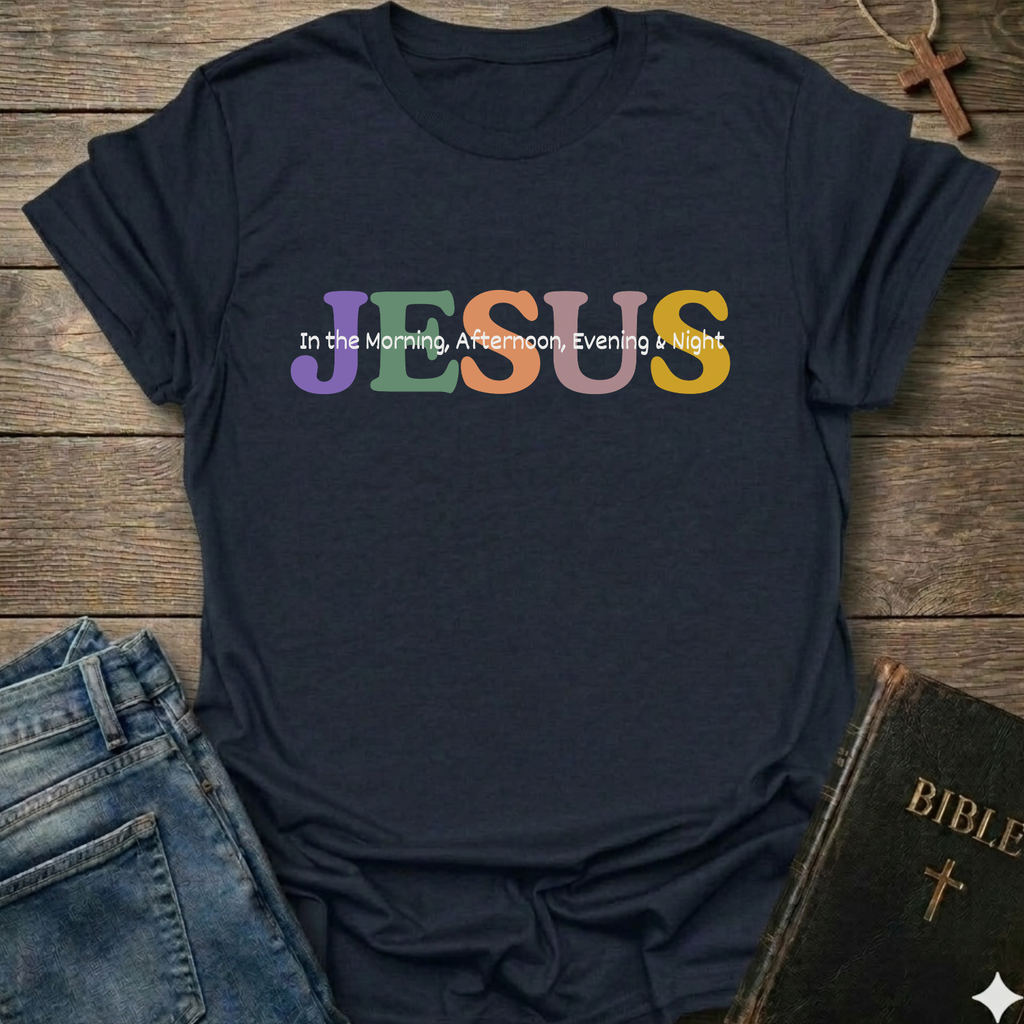 A navy blue graphic t-shirt laid flat on a rustic wooden background, styled with blue jeans, a Bible, and a cross necklace. The shirt features the word 'JESUS' in large, colorful pastel letters (purple, green, orange, yellow) with the phrase 'In the Morning, Afternoon, Evening & Night' printed in white script on top of it.