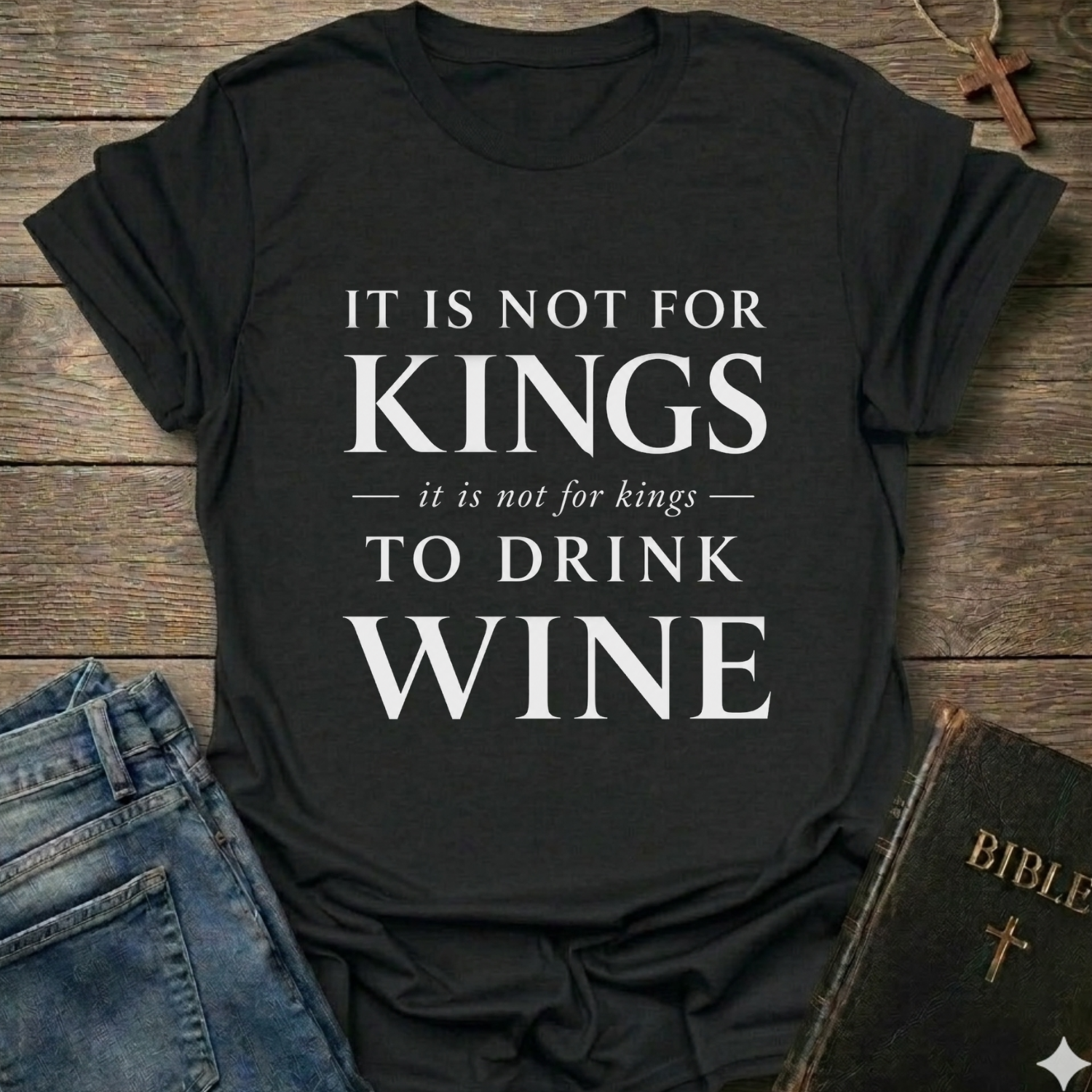 A black graphic t-shirt with the text 'IT IS NOT FOR KINGS it is not for kings TO DRINK WINE' in white, styled in a flat-lay on a rustic wooden background with blue jeans and a Bible.