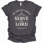 A dark heather gray short-sleeve t-shirt with a cream-colored graphic print that reads 'ME AND MY HOUSE SERVE THE LORD - JOSHUA 24:15' with a house silhouette and cross.