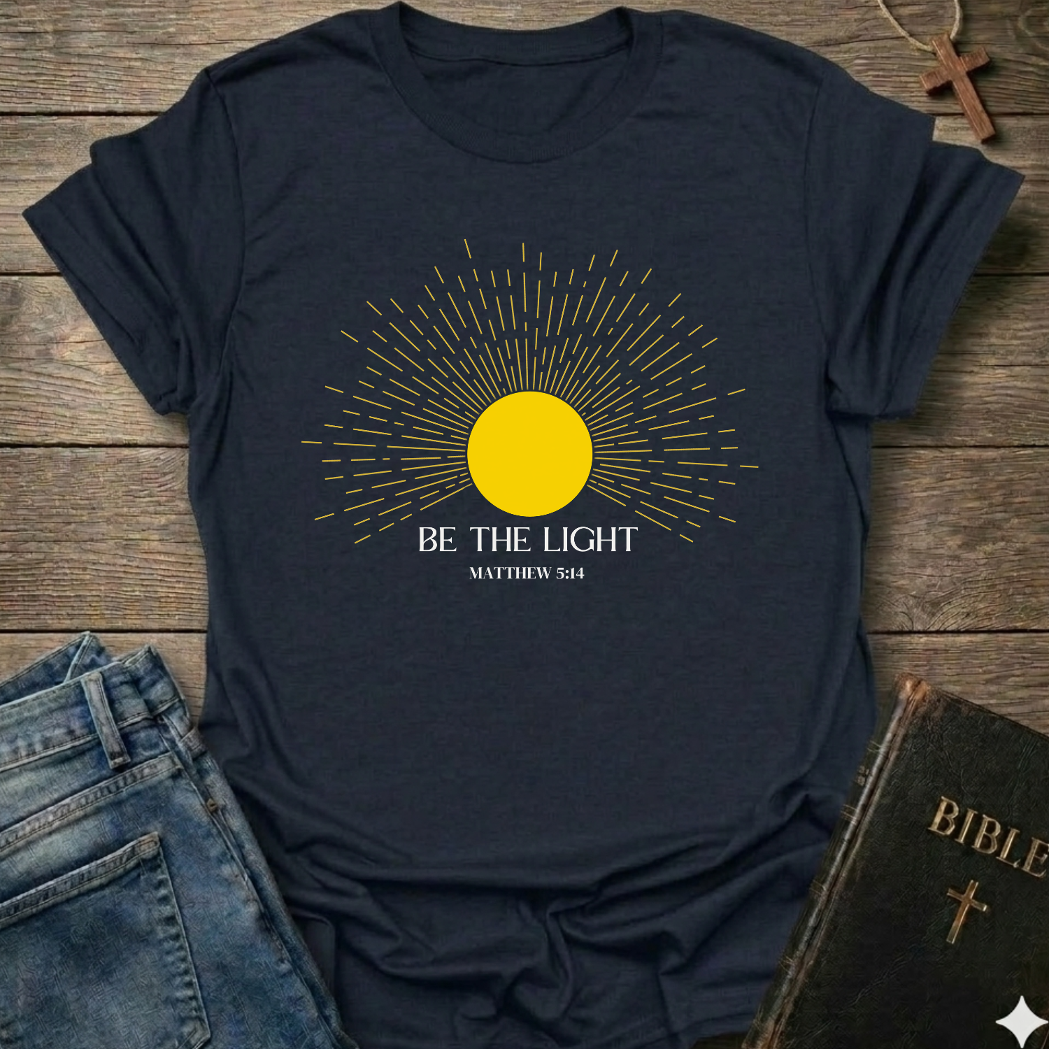Navy blue t-shirt with yellow sun graphic and 'Be the Light' text on a wooden surface with jeans and a Bible. Be the Light Christian T-Shirt Matthew 5:14 bold faith apparel Faith Forward