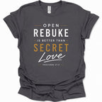 A dark heather gray graphic t-shirt featuring the text 'OPEN REBUKE IS BETTER THAN SECRET Love' and 'PROVERBS 27:5' in white and gold lettering, displayed on a clean white background.