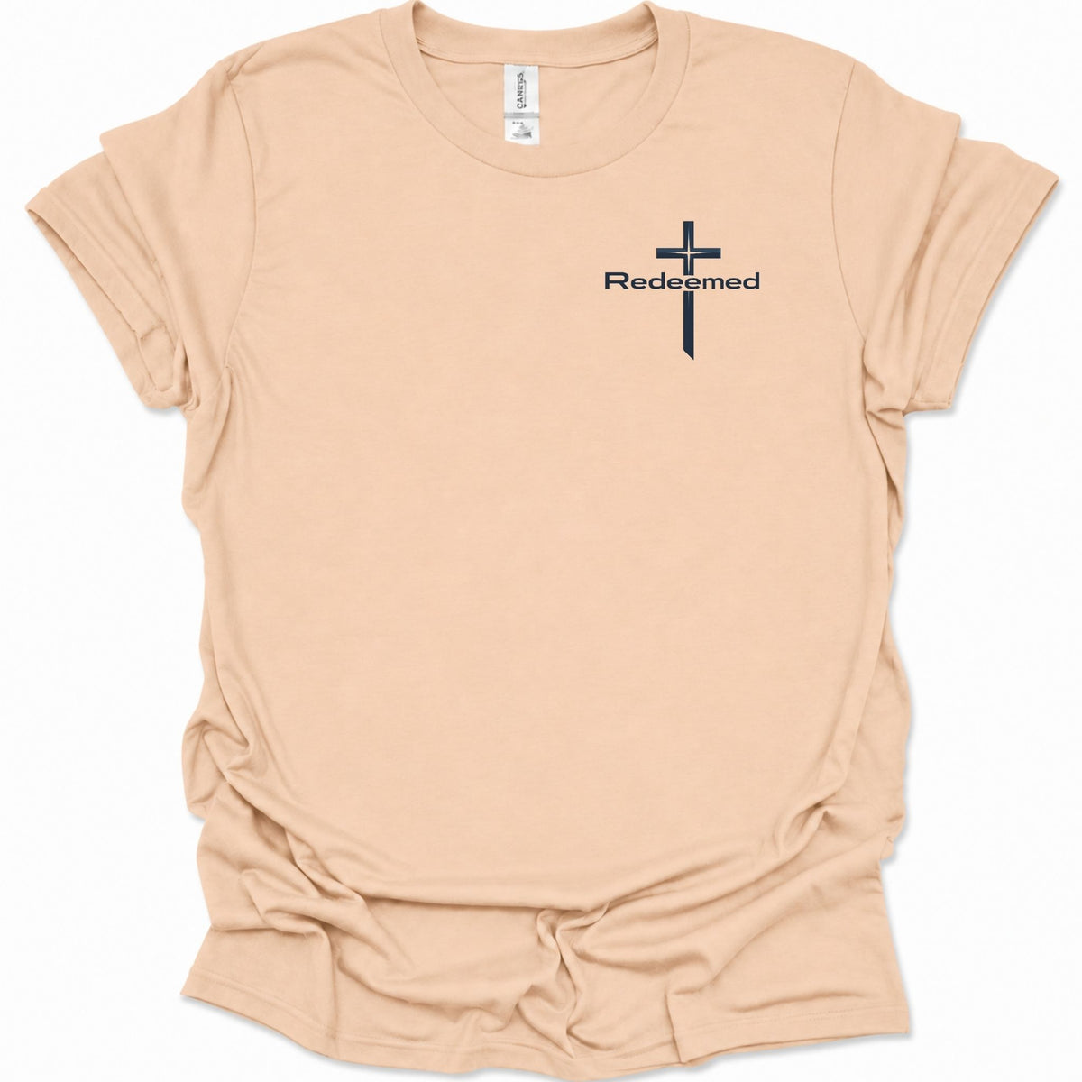 A light natural-colored short-sleeve t-shirt with a black cross graphic and the word 'Redeemed' printed on the left chest, displayed on a clean white background.