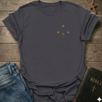 Dark gray t-shirt with Three Dots and a Cross Minimal Christian T-Shirt conversation starter faith apparel on a wooden surface with jeans and a Bible.