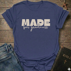 A purple  t-shirt with the text 'MADE for greatness' based on 2 Corinthians 5 verse 17 printed on the front, styled in a flat-lay arrangement with blue jeans and a Bible on a rustic wooden background.