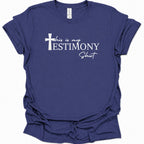 A purple blue short-sleeve t-shirt with a white graphic print on the front that reads 'this is my TESTIMONY Shit' alongside a cross icon, displayed against a clean white background.