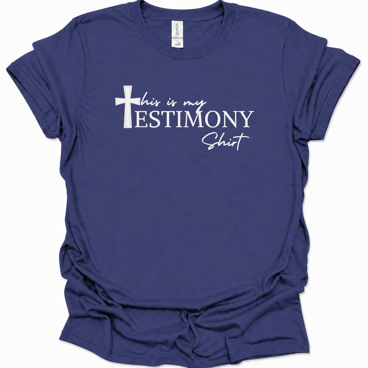 A purple blue short-sleeve t-shirt with a white graphic print on the front that reads 'this is my TESTIMONY Shit' alongside a cross icon, displayed against a clean white background.