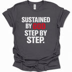 A dark heather gray short-sleeve t-shirt with a graphic text print reading 'SUSTAINED BY GOD STEP BY STEP.' in white and red lettering, displayed on a clean white background.