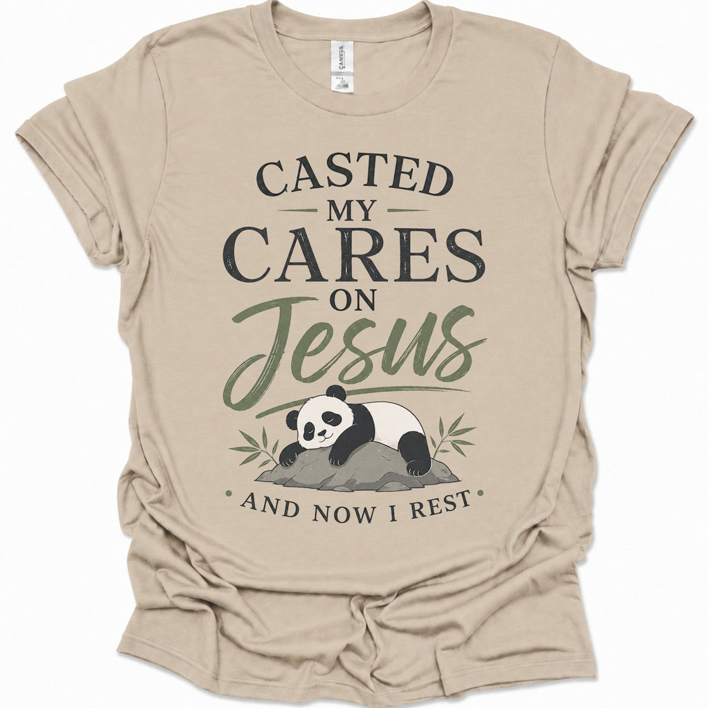 A sand colored short-sleeve t-shirt with a graphic print that reads 'CASTED MY CARES ON Jesus AND NOW I REST' in black and olive green lettering, accompanied by an illustration of a sleeping panda resting on a rock with bamboo leaves.