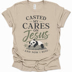 A sand colored short-sleeve t-shirt with a graphic print that reads 'CASTED MY CARES ON Jesus AND NOW I REST' in black and olive green lettering, accompanied by an illustration of a sleeping panda resting on a rock with bamboo leaves.
