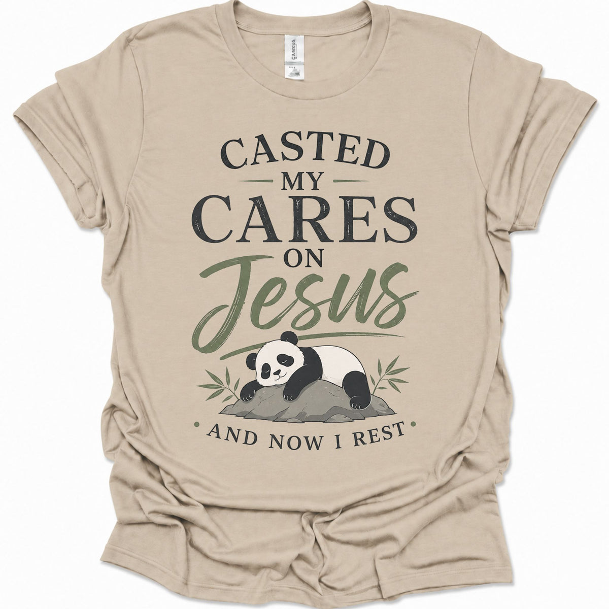 A sand colored short-sleeve t-shirt with a graphic print that reads 'CASTED MY CARES ON Jesus AND NOW I REST' in black and olive green lettering, accompanied by an illustration of a sleeping panda resting on a rock with bamboo leaves.