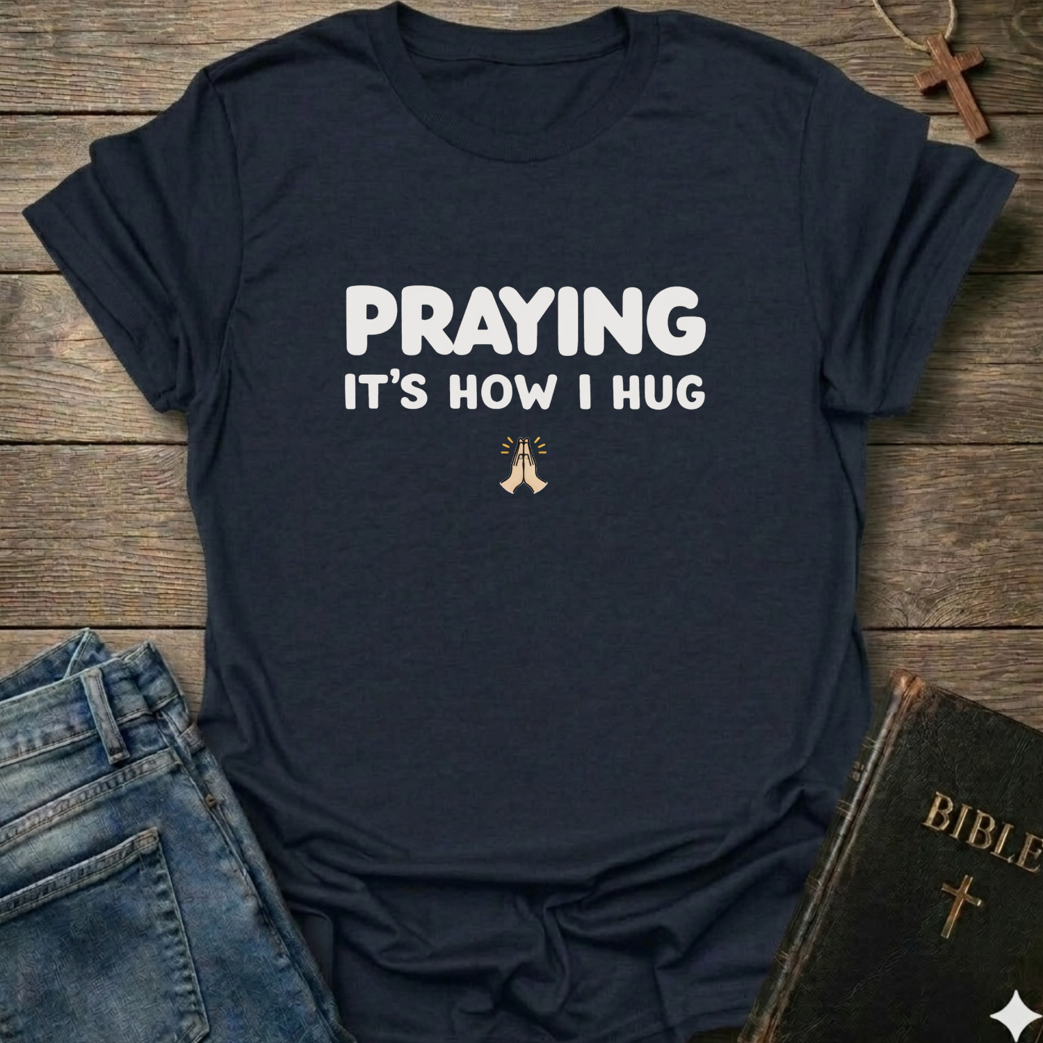 Black t-shirt with 'PRAYING IT'S HOW I HUG' text Christian faith apparel relatable scripture tee conversation starter on a wooden surface with jeans and a Bible.