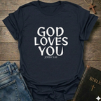 Navy blue t-shirt with 'GOD LOVES YOU' John 3:16 text Christian bold faith apparel Faith Forward on a wooden surface with jeans and a Bible.