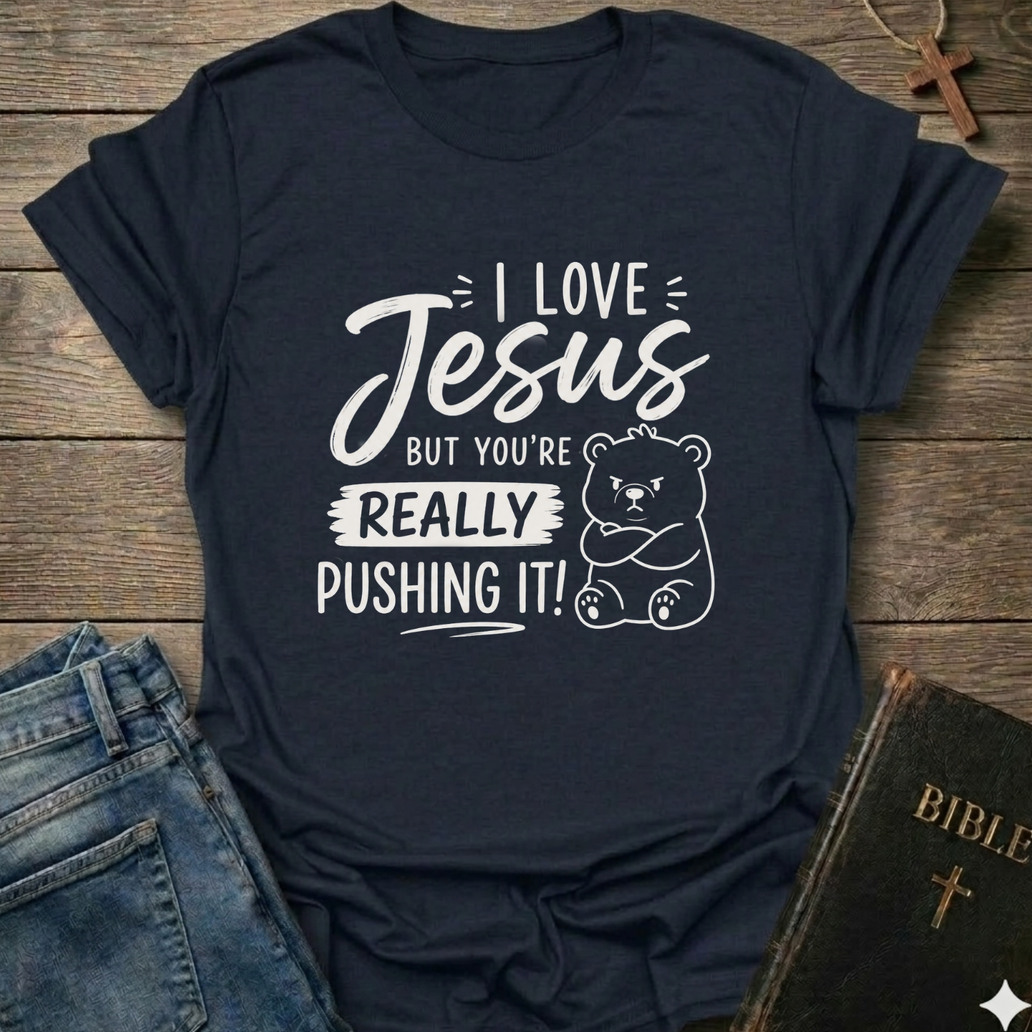 A navy blue graphic t-shirt laid flat on a rustic wooden background, styled with blue jeans, a Bible, and a wooden cross. The t-shirt features a white printed graphic that reads 'I LOVE Jesus BUT YOU'RE REALLY PUSHING IT!' alongside a cartoon bear illustration.