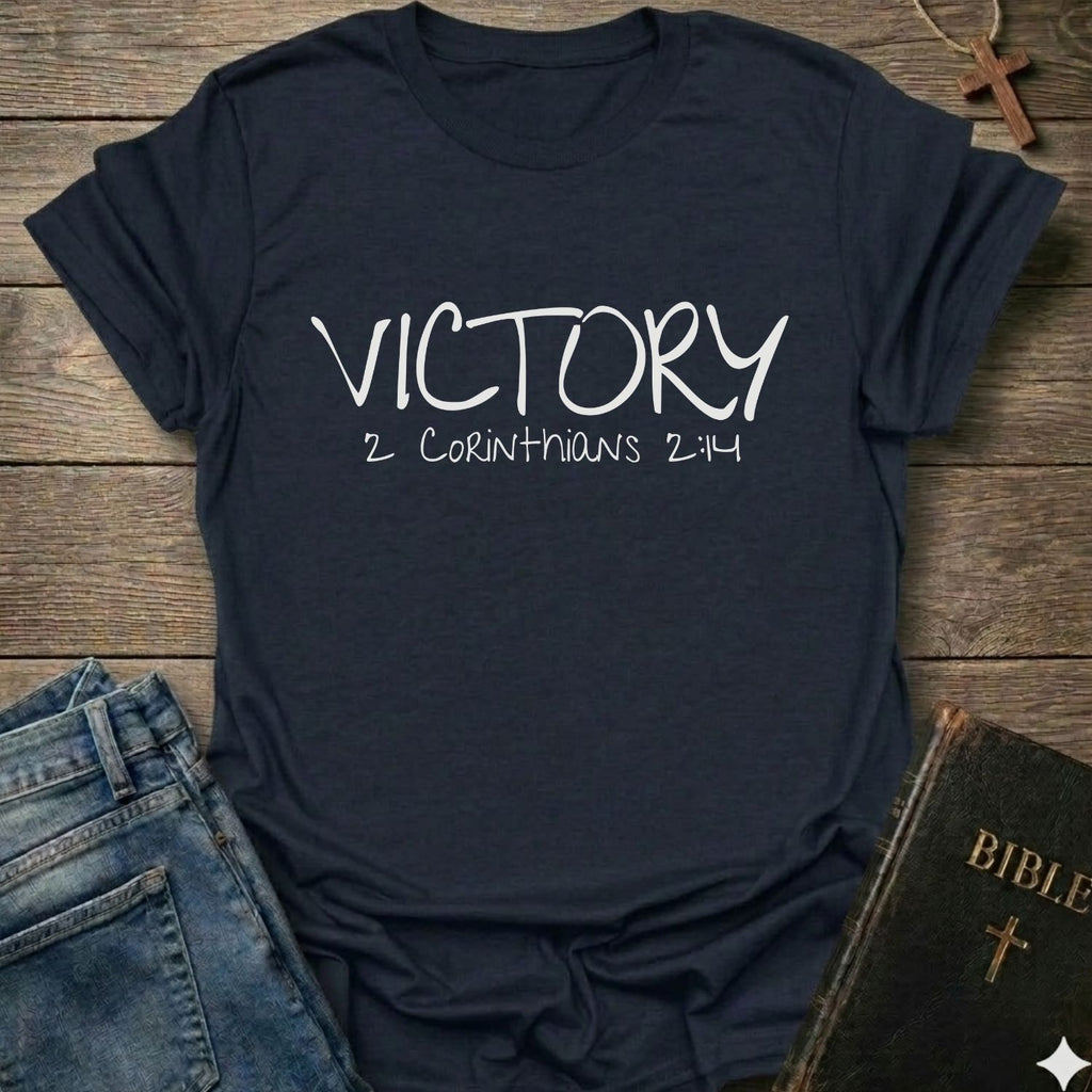 A navy blue graphic t-shirt with 'VICTORY 2 CORINTHIANS 2:14' printed in white script, styled in a flat-lay on a wooden background with jeans, a Bible, and a cross necklace.