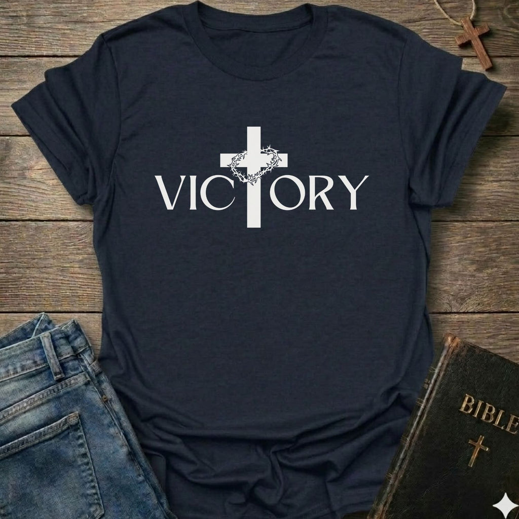 A navy blue short-sleeve t-shirt with a white graphic print of a cross and the word 'VICTORY' across the chest, styled in a flat-lay arrangement with blue jeans, a Bible, and a wooden cross on a rustic wood background.