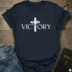 A navy blue short-sleeve t-shirt with a white graphic print of a cross and the word 'VICTORY' across the chest, styled in a flat-lay arrangement with blue jeans, a Bible, and a wooden cross on a rustic wood background.