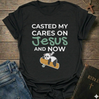 Black t-shirt Casted My Cares on Jesus Christian relatable faith apparel conversation starter with a panda graphic and text on a wooden surface with jeans and a Bible.