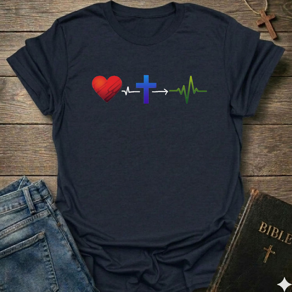 A dark navy blue t-shirt laid flat on a rustic wooden background, styled with blue jeans, a Bible, and a wooden cross necklace. The t-shirt features a graphic design of a red heart, a blue cross, and a green heartbeat line connected by white arrows.
