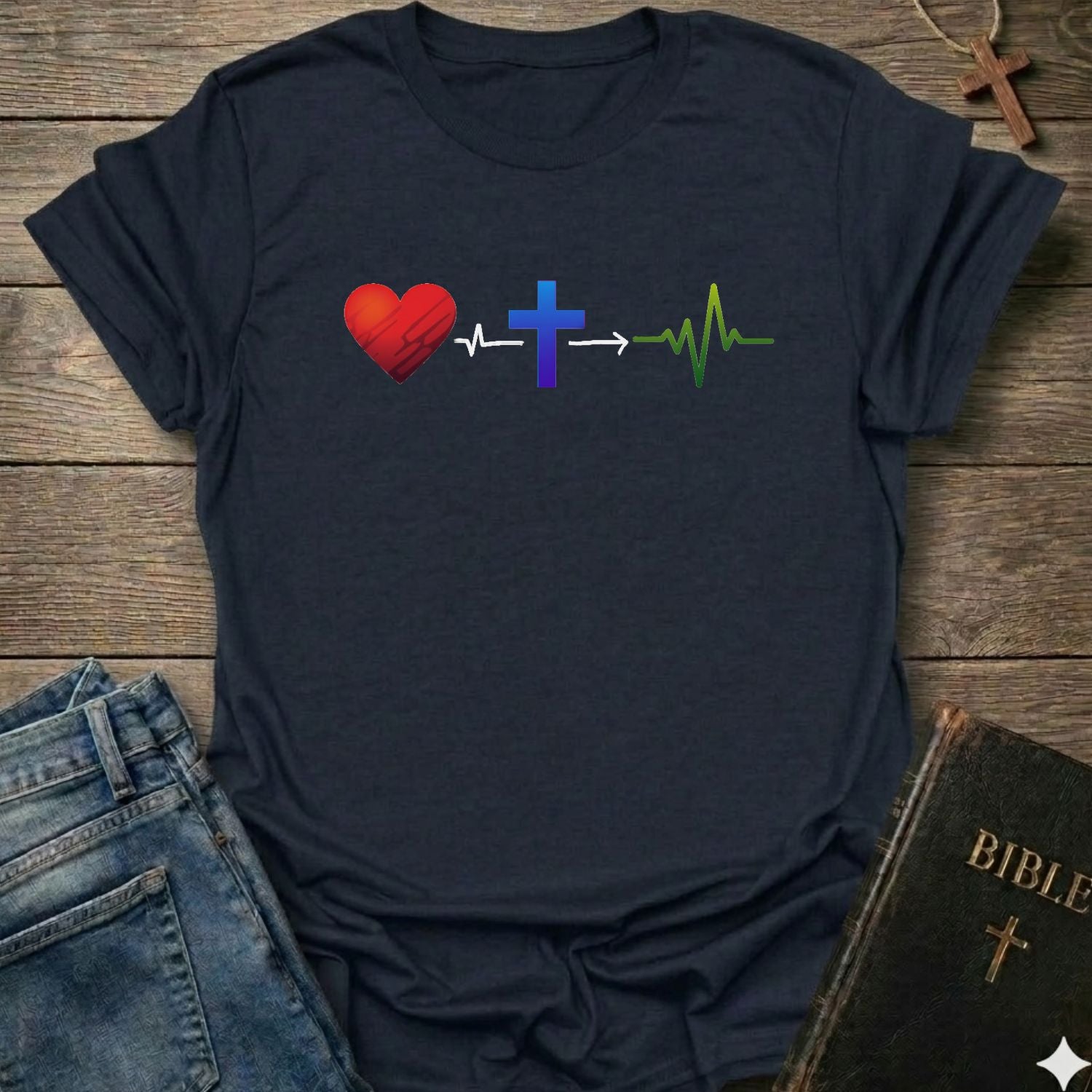 A dark navy blue t-shirt laid flat on a rustic wooden background, styled with blue jeans, a Bible, and a wooden cross necklace. The t-shirt features a graphic design of a red heart, a blue cross, and a green heartbeat line connected by white arrows.