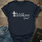 Navy blue t-shirt with 'This is my testimony shirt' text and cross minimal faith apparel conversation starter. It is on a wooden surface with jeans and a Bible.