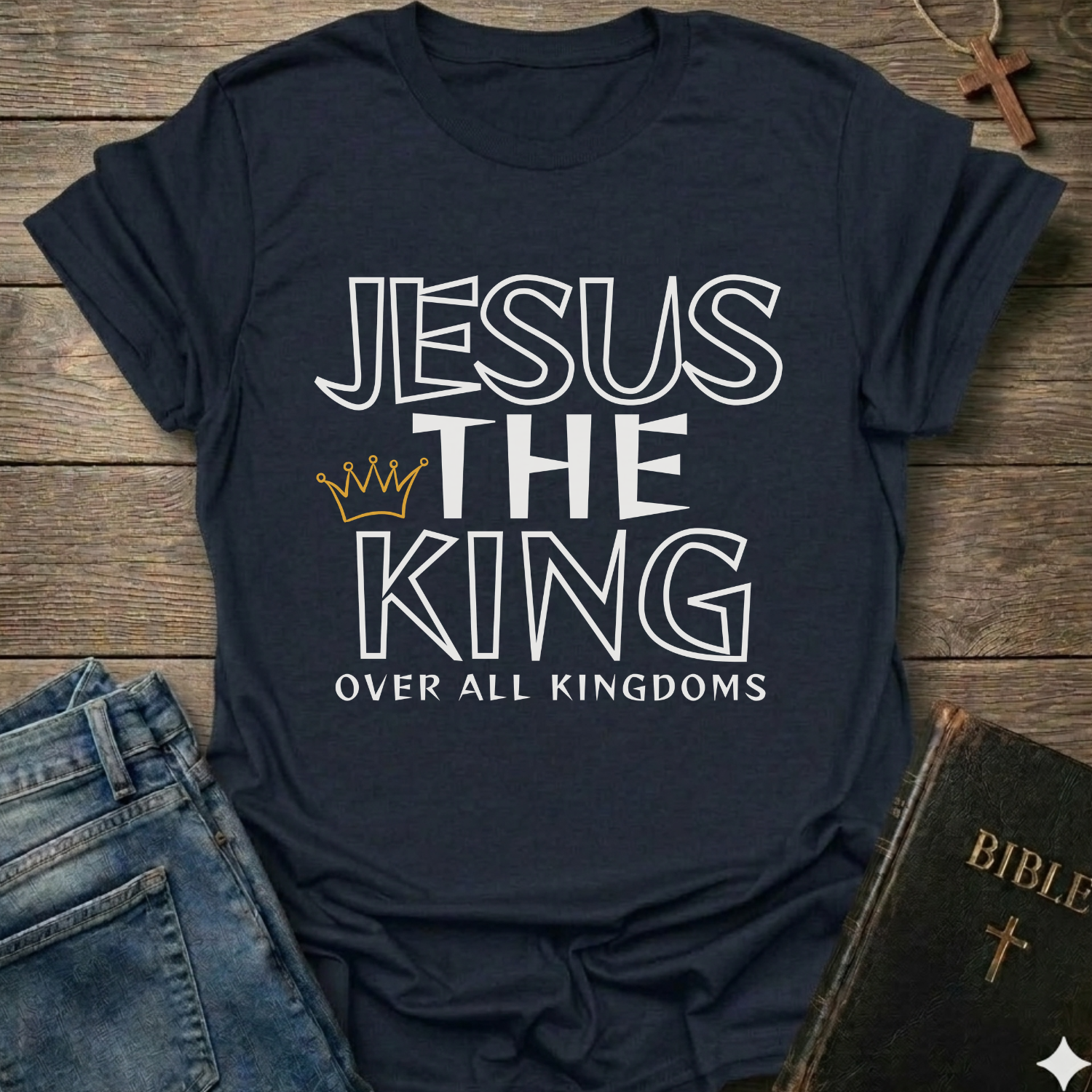 Navy blue t-shirt with text 'Jesus The King Over All Kingdoms'  and crown Christian bold faith apparel Faith Forward text on a wooden surface with jeans and a Bible.