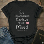 A black graphic t-shirt laid flat on a rustic wooden surface, styled with blue jeans, a Bible, and a wooden cross. The t-shirt features the white text 'Be Transformed Renew Your Mind Romans 12:2' with a small red brain graphic.