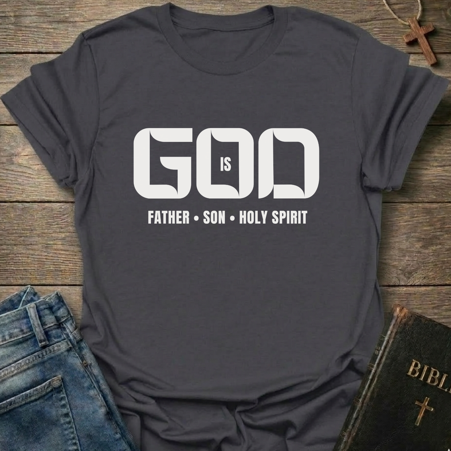 A dark heather gray short-sleeve graphic T-shirt laid flat on a rustic wooden surface, styled with blue jeans, a Bible, and a cross. The shirt has large white text reading 'GOD IS' with 'FATHER • SON • HOLY SPIRIT' underneath.