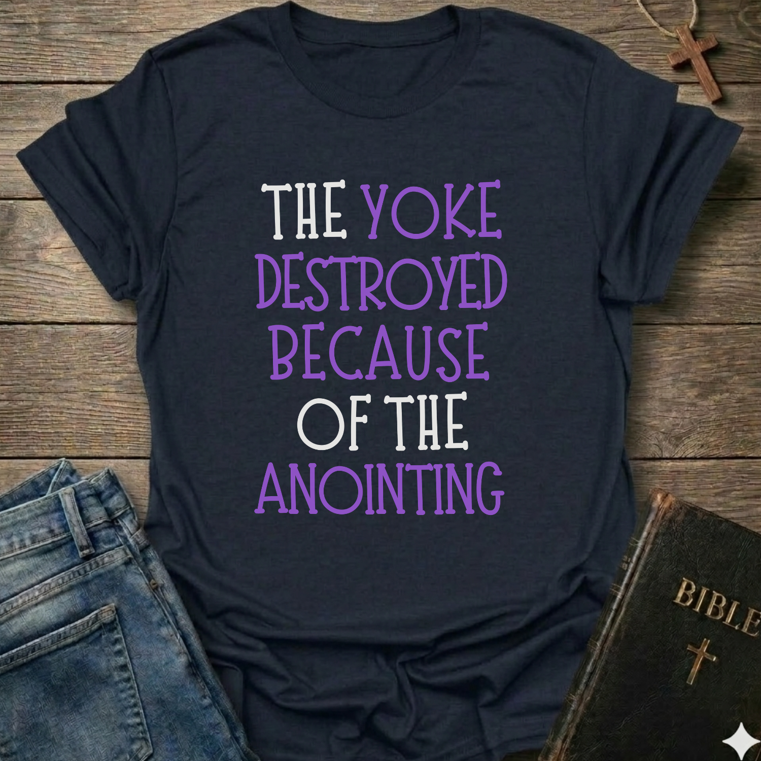Black t-shirt with text 'The yoke destroyed because of the anointing' Isaiah 10:27 faith apparel conversation starter on a wooden surface with jeans and a Bible.