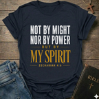A navy blue graphic t-shirt laid flat on a rustic wooden background, styled with blue jeans and a Bible. The t-shirt features the text 'NOT BY MIGHT NOR BY POWER BUT BY MY SPIRIT ZECHARIAH 4:6' in white and gold lettering.