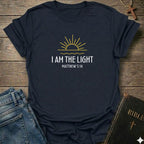 A navy blue graphic t-shirt laid flat on a rustic wooden background, styled with a pair of blue jeans, a Bible, and a wooden cross. The t-shirt features a yellow sun graphic and the text 'I AM THE LIGHT MATTHEW 5:14' in white.