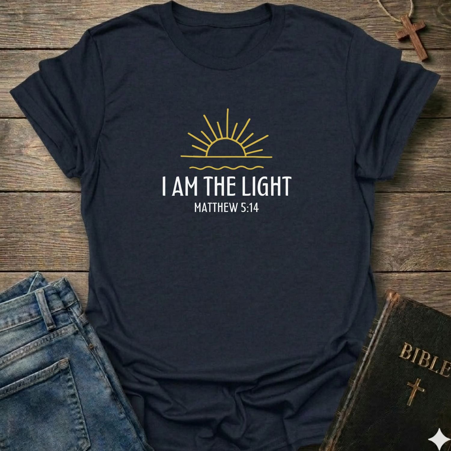 A navy blue graphic t-shirt laid flat on a rustic wooden background, styled with a pair of blue jeans, a Bible, and a wooden cross. The t-shirt features a yellow sun graphic and the text 'I AM THE LIGHT MATTHEW 5:14' in white.
