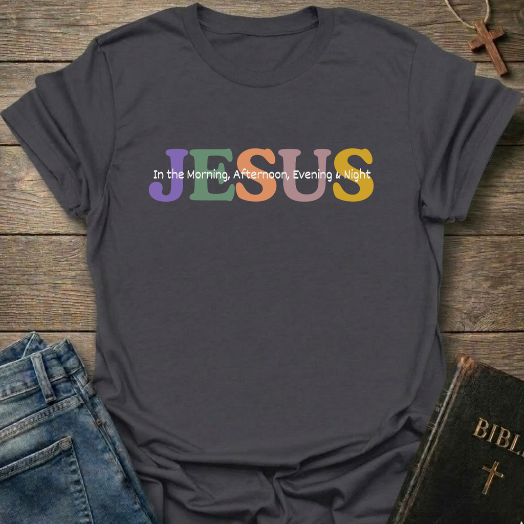 A dark charcoal gray t-shirt laid flat on a rustic wooden background, styled with blue jeans, a Bible, and a cross necklace. The shirt features the word 'JESUS' in large, colorful pastel letters (purple, green, orange, yellow) with the phrase 'In the Morning, Afternoon, Evening & Night' printed in white script on top of it.
