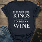 A navy blue graphic t-shirt with the text 'IT IS NOT FOR KINGS it is not for kings TO DRINK WINE' in white, styled in a flat-lay on a wooden background with jeans, a Bible, and a cross.