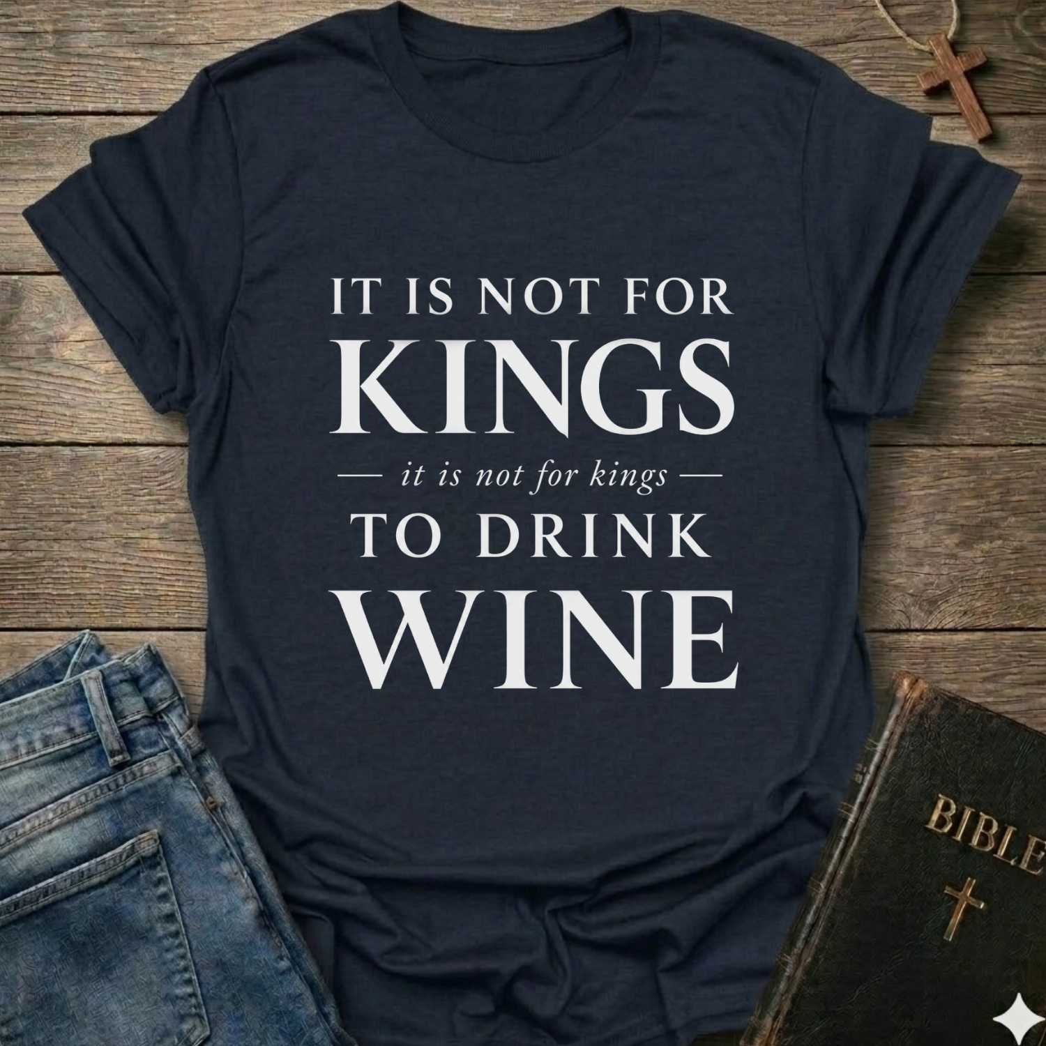 A navy blue graphic t-shirt with the text 'IT IS NOT FOR KINGS it is not for kings TO DRINK WINE' in white, styled in a flat-lay on a wooden background with jeans, a Bible, and a cross.
