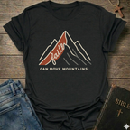 Black Christian t-shirt with 'Faith can move mountains' graphic Matthew 17:20 faith apparel conversation starter on a wooden surface with jeans and a Bible. 