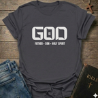 Gray t-shirt with 'GOD IS FATHER SON HOLY SPIRIT' text on a wooden surface with jeans and a Bible.