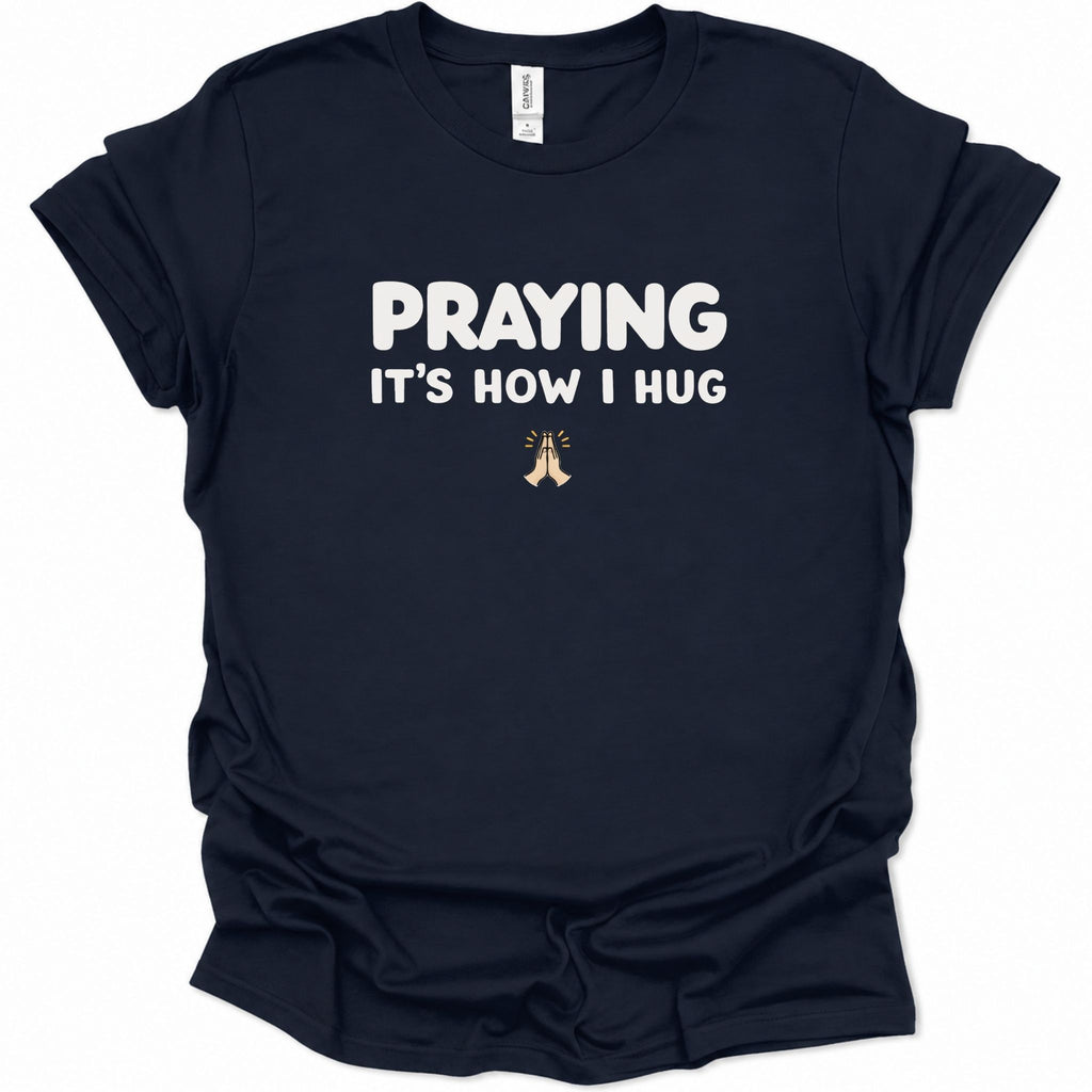 A navy blue short-sleeve t-shirt with white text reading 'PRAYING IT'S HOW I HUG' and a small praying hands graphic below, shown on a plain white background.