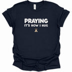 A navy blue short-sleeve t-shirt with white text reading 'PRAYING IT'S HOW I HUG' and a small praying hands graphic below, shown on a plain white background.