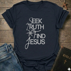 Navy blue t-shirt with 'Seek Truth Find Jesus' text on a wooden surface with jeans and a Bible. Bold typography faith apparel conversation starter.