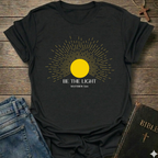 Black t-shirt with yellow sun graphic and 'Be the Light' text on a wooden surface with jeans and a Bible. Be the Light Christian T-Shirt Matthew 5:14 bold faith apparel Faith Forward