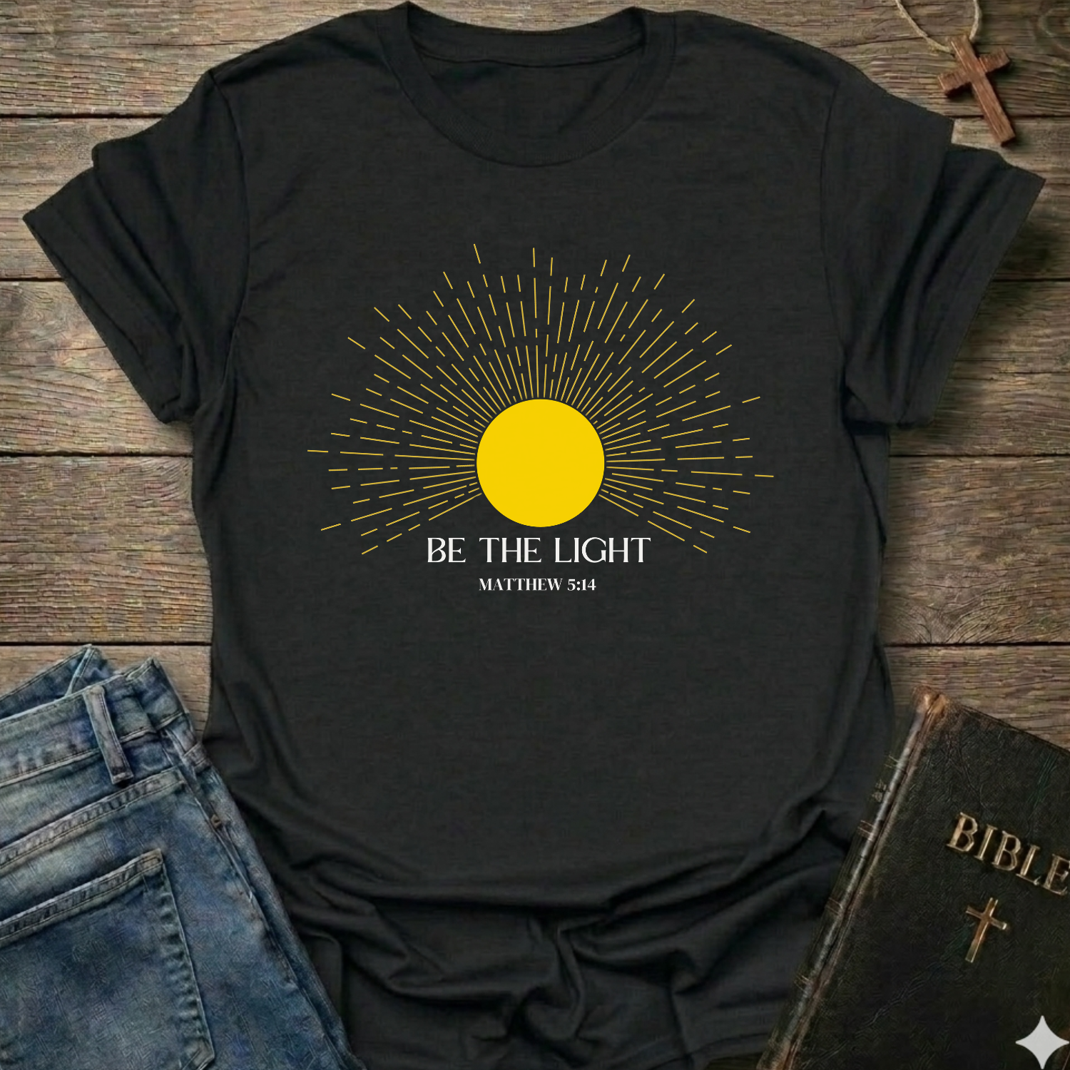 Black t-shirt with yellow sun graphic and 'Be the Light' text on a wooden surface with jeans and a Bible. Be the Light Christian T-Shirt Matthew 5:14 bold faith apparel Faith Forward