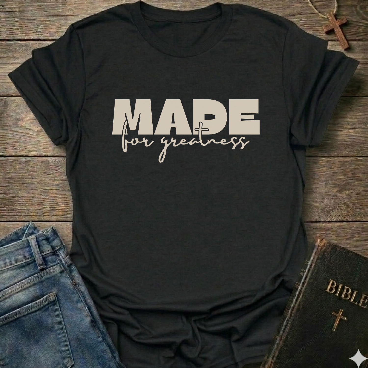 A black short-sleeve T-shirt laid flat on a rustic wooden surface, with the text 'MADE for greatness' based on 2 Corinthians 5 verse 17 printed on the front, styled in a flat-lay arrangement with blue jeans and a Bible on a rustic wooden background.