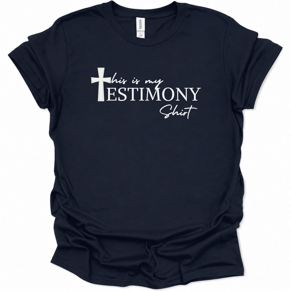 A navy blue short-sleeve t-shirt with a white graphic text print reading 'this is my TESTIMONY Shit' alongside a white cross icon, displayed on a clean white background.