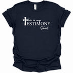 A navy blue short-sleeve t-shirt with a white graphic text print reading 'this is my TESTIMONY Shit' alongside a white cross icon, displayed on a clean white background.