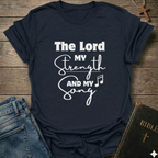 A navy blue graphic t-shirt with white text reading 'The Lord MY Strength AND MY Song' is laid flat on a rustic wooden background, styled with blue jeans on the left and a Bible with a cross on the right.