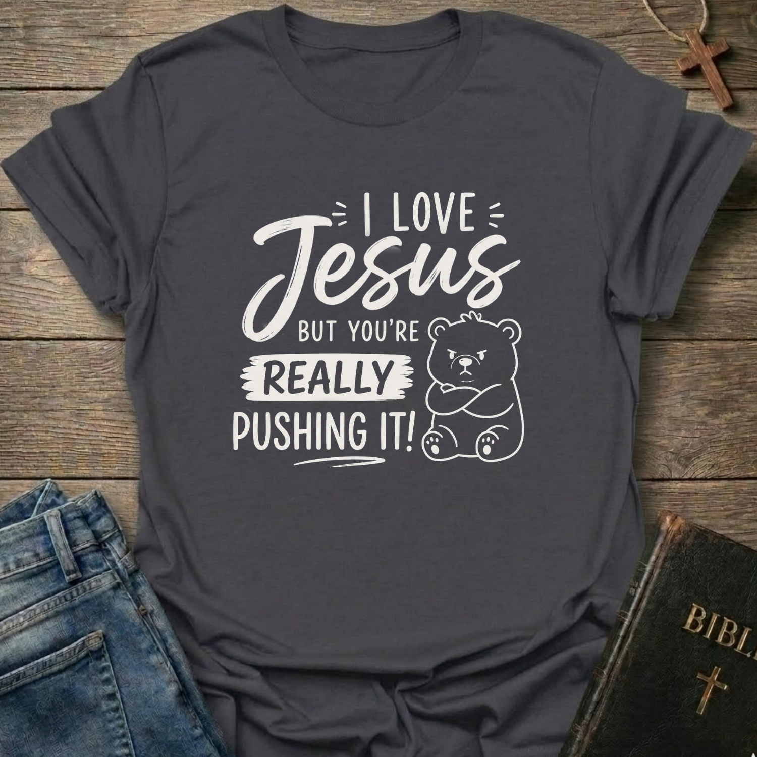 A dark heather  gray graphic t-shirt laid flat on a rustic wooden background, styled with blue jeans, a Bible, and a wooden cross. The t-shirt features a white printed graphic that reads 'I LOVE Jesus BUT YOU'RE REALLY PUSHING IT!' alongside a cute cartoon bear.