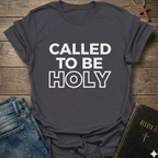 Gray t-shirt with 'Called to be Holy' Christian T-Shirt bold faith apparel 1 Peter 1:15 conversation starter text on a wooden surface with jeans and a Bible.