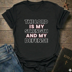 Black t-shirt with 'The Lord is my strength and my defense' Exodus 15:2 bold faith apparel Faith Forward on a wooden surface with jeans and a Bible.