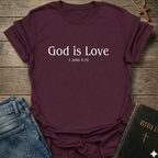 Maroon t-shirt with 'God is Love'  Christian T-Shirt 1 John 4:15 elegant faith apparel Faith Forward text. T-shirt on a wooden surface with jeans and a Bible.