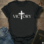 A black short-sleeve T-shirt laid flat on a rustic wooden surface, featuring a white cross graphic with a small floral detail and the word 'VICTORY' printed across the chest. Blue jeans and a dark Bible are partially visible as styling props.
